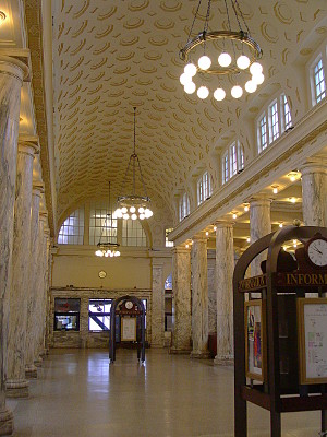 [This hallway contains wooden information kiosks with clocks atop them. The ceiling is curved and has circular depressions. Three chandeliers with globe bulbs hang from the ceilings. The marble columns are topped by a row of windows before reaching the curved ceiling.]